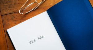 a pair of glasses and a book with a dark blue back cover and the last page showing the words The End sit atop a brown wood desk.